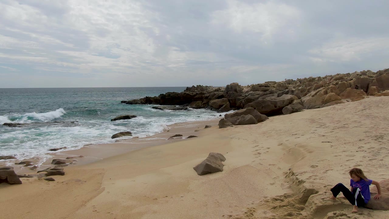 Mid-clip frame at Shipwrecks East Cape BCS Mexico with a wave breaking near the rocks, sandy foreground, cloudy horizon, and a person in purple shifting on the lower right slope.