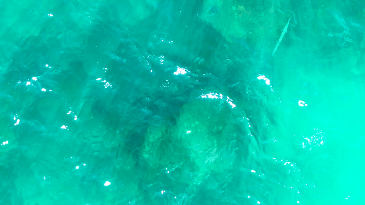 Overhead view of turquoise water at Buzzards Beach East Cape BCS Mexico with dark underwater rocks, rippled surface glare, and no visible people.
