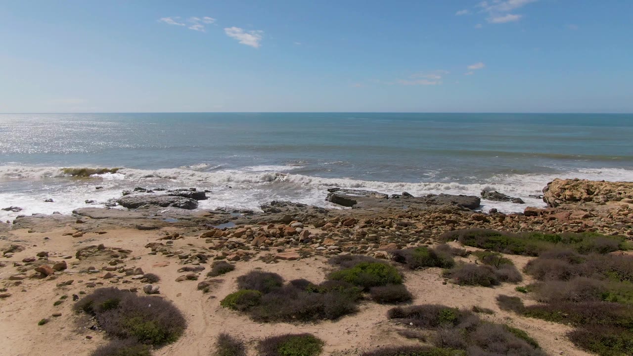 At Buzzards Beach East Cape BCS Mexico, surf spreads across dark rock shelves while tan sand, scattered stones, and low shrubs fill the lower frame, with no visible people.