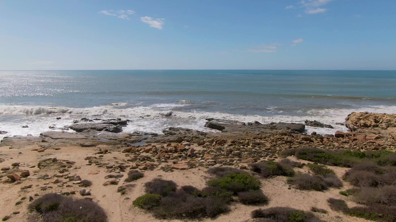 At Buzzards Beach East Cape BCS Mexico, a band of white foam pushes onto the rock shelf, with dry sand, small boulders, and green brush in the foreground and no visible people.