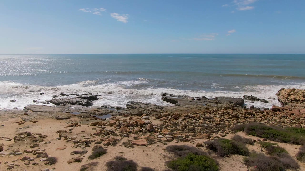 At Buzzards Beach East Cape BCS Mexico, bright ocean glare sits left of frame as waves break across the rocks and a field of beach stones leads into low scrub, with no visible people.