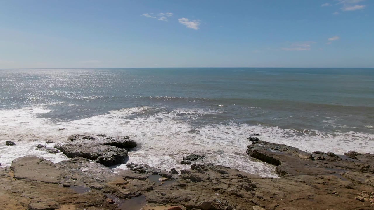 At Buzzards Beach in Mexico, foamy water breaks around flat tan and dark rocks while sunlight reflects off the ocean, with no visible people.