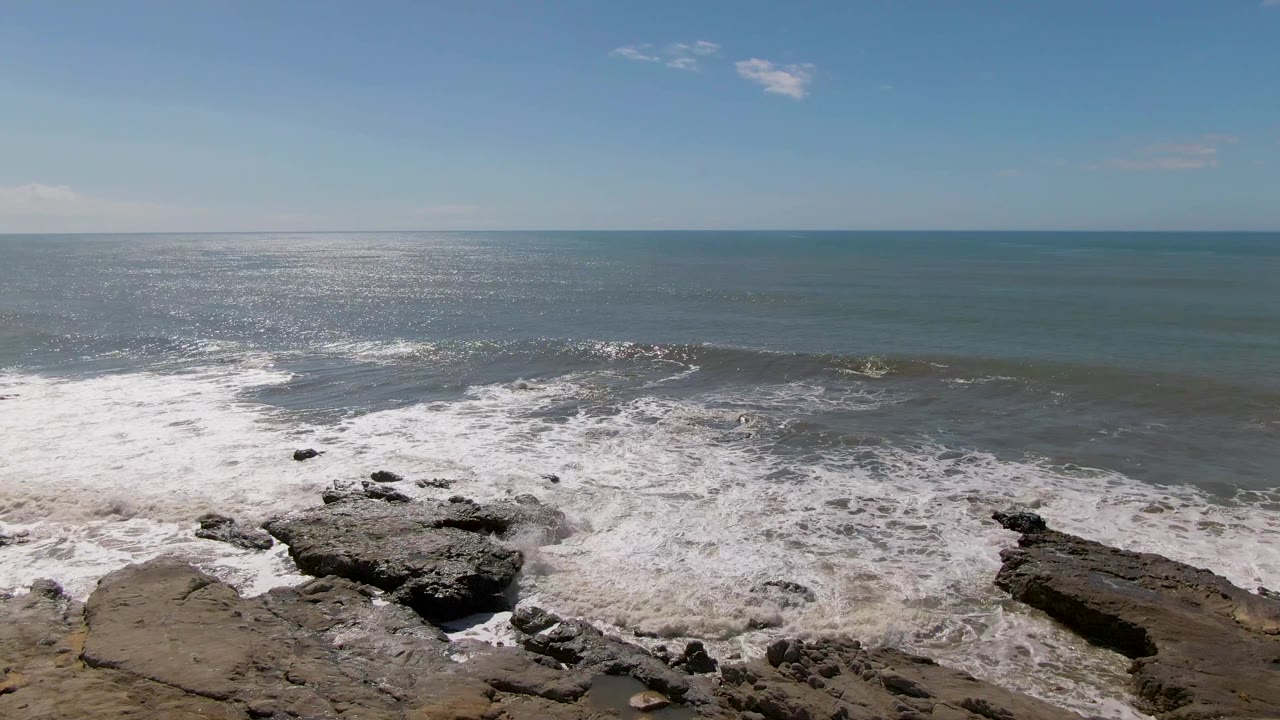 At Buzzards Beach in Mexico, strong sunlight glitters on the left side of the ocean while a wide band of surf breaks over rocks below, with no visible people.