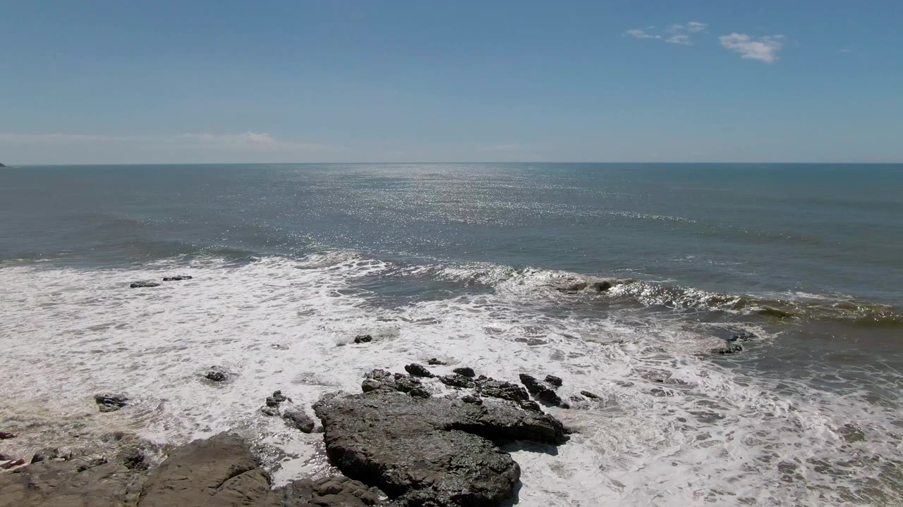 At Buzzards Beach in East Cape BCS Mexico, a broad band of whitewater spreads toward foreground rocks while the open ocean and blue sky fill the background with no visible people.