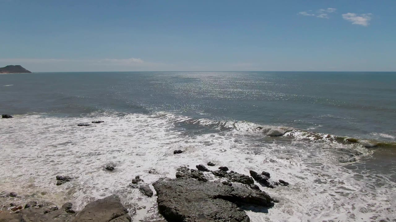 At Buzzards Beach in East Cape BCS Mexico, waves break across a rocky shoreline while a distant headland sits near the horizon and no visible people appear in frame.