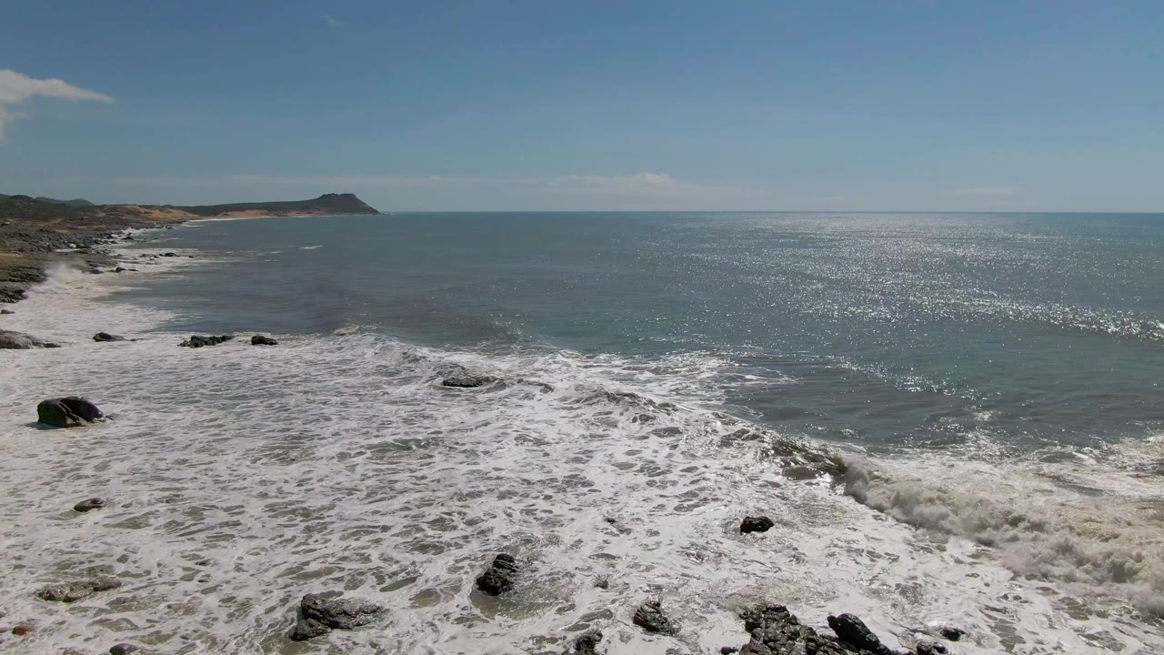 Opening aerial view of Buzzards Beach East Cape BCS Mexico with white surf washing over dark rocks, open ocean to the right, coastal scrub to the left, and no visible people.