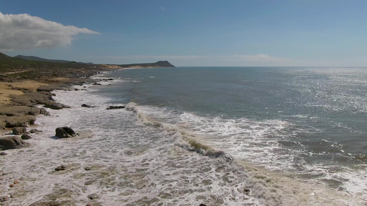 Mid-clip aerial view at Buzzards Beach East Cape BCS Mexico with the surf zone lower in frame, calm open water across the right side, scrubby coastline at left, and no visible people.