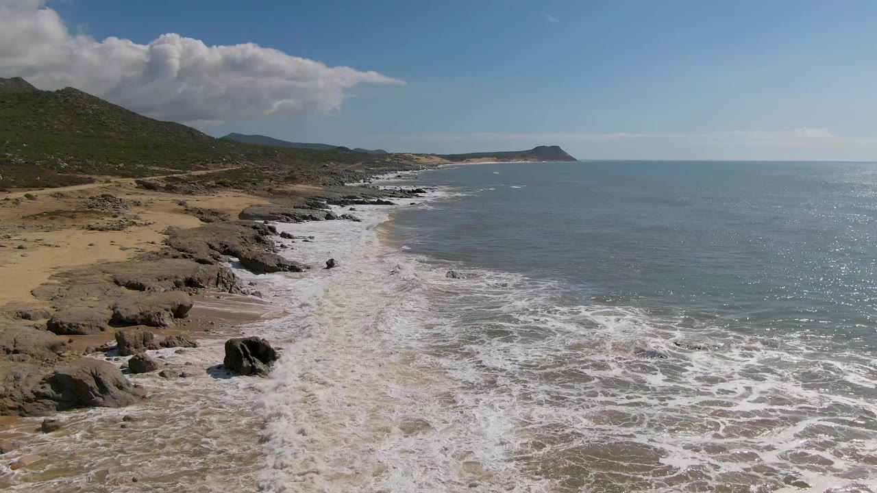 Aerial coastal view at Buzzards Beach in Mexico with a large foreground rock, whitewater spreading over wet sand, scrubby hills on the left, ocean horizon on the right, and no visible people.