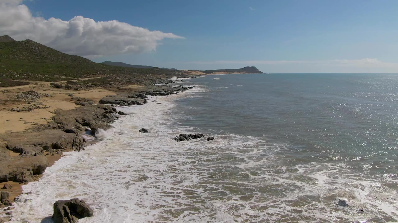 Aerial view at Buzzards Beach East Cape BCS Mexico showing white surf washing over rocks beside tan sand, green hills to the left, open ocean to the right, and no visible people.