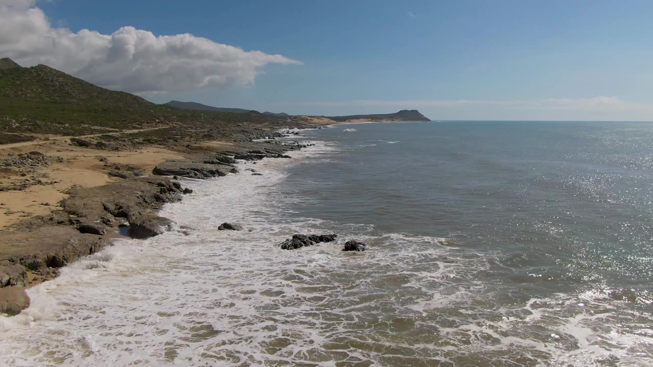 At 0:01, Buzzards Beach surf spreads farther across the near rocks, with sandy beach at left, hills behind the shoreline, bright ocean glare at right, and no visible people.