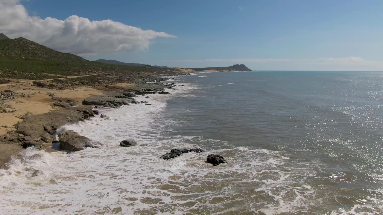 At 0:02, the rocky shoreline remains prominent at Buzzards Beach, with white water breaking across the lower frame, a distant coastal point ahead, and no visible people.