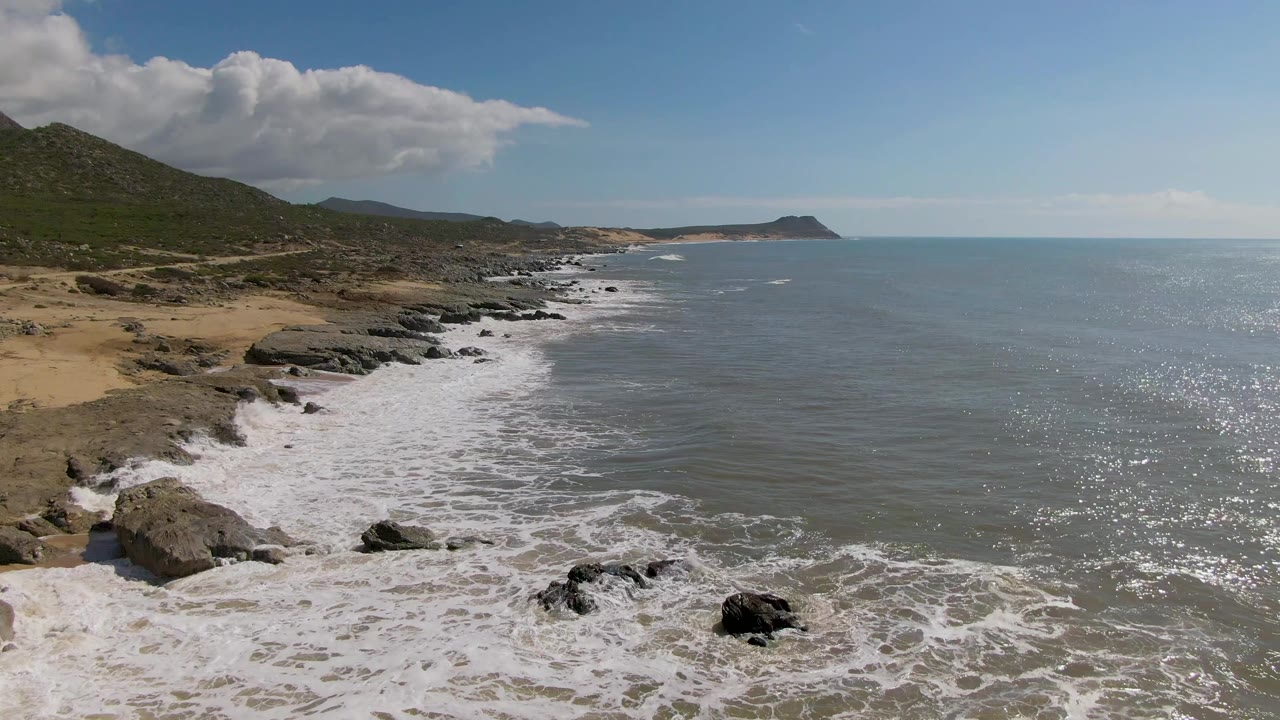 Coastal waves roll toward dark rocks along a sandy and hilly shore.