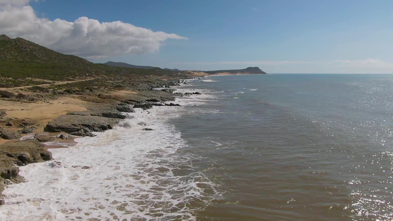 Aerial view of waves breaking along a rocky beach with hills and open ocean.