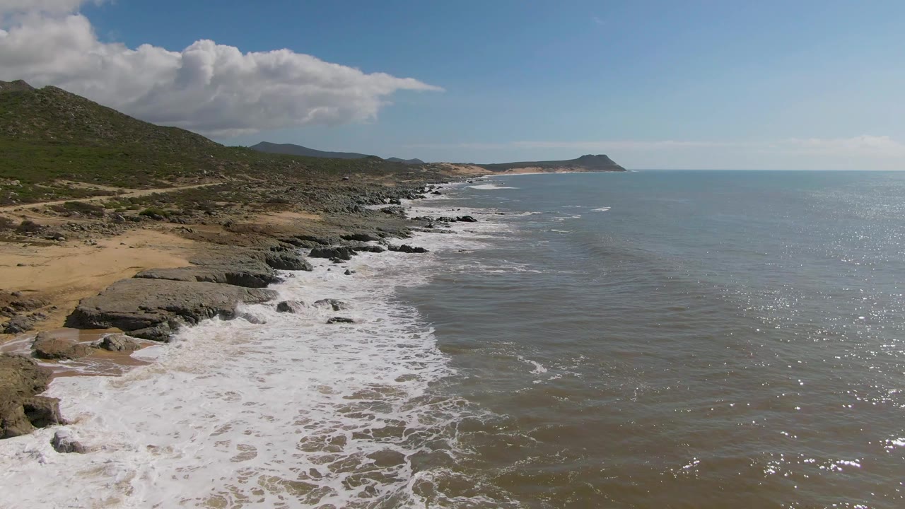 Buzzards Beach East Cape BCS Mexico coastline seen from above as foamy water pushes farther over the rocks, with sandy beach on the left, ocean to the right, and no visible people.