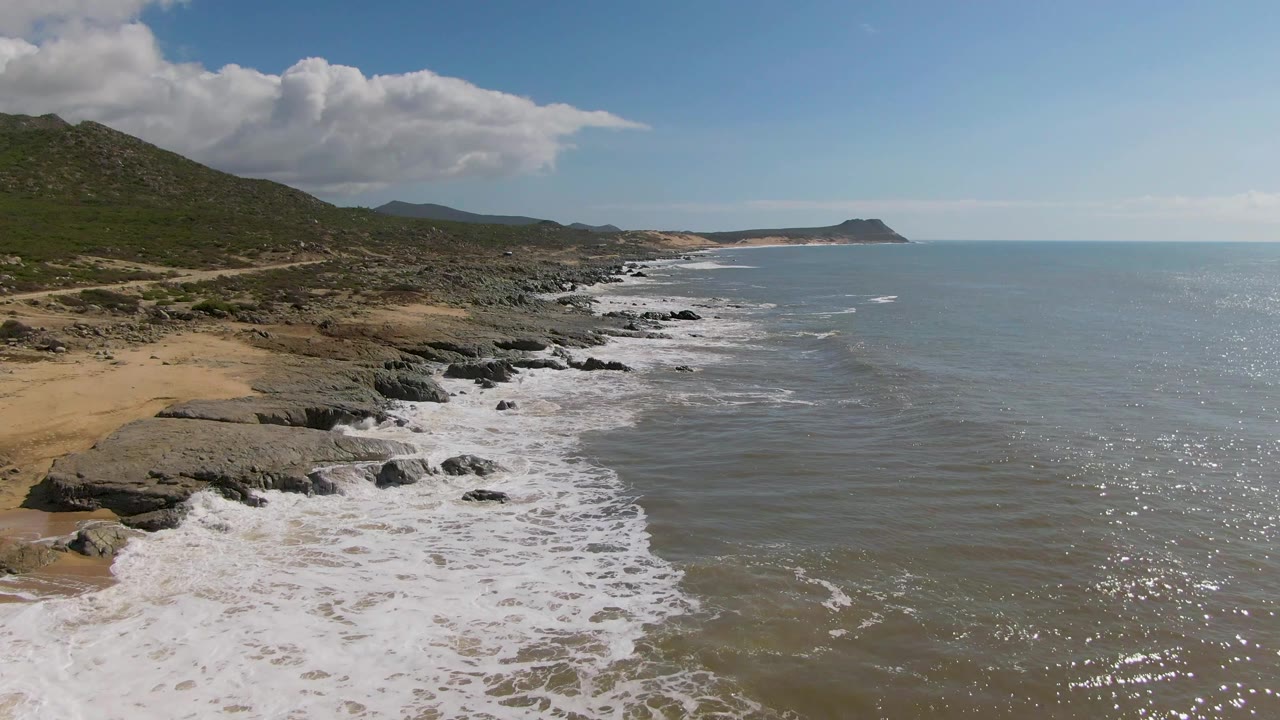 Aerial coastal frame from Buzzards Beach East Cape BCS Mexico with white water stretching along the rocky shore, brown-green hills inland, a distant headland on the horizon, and no visible people.