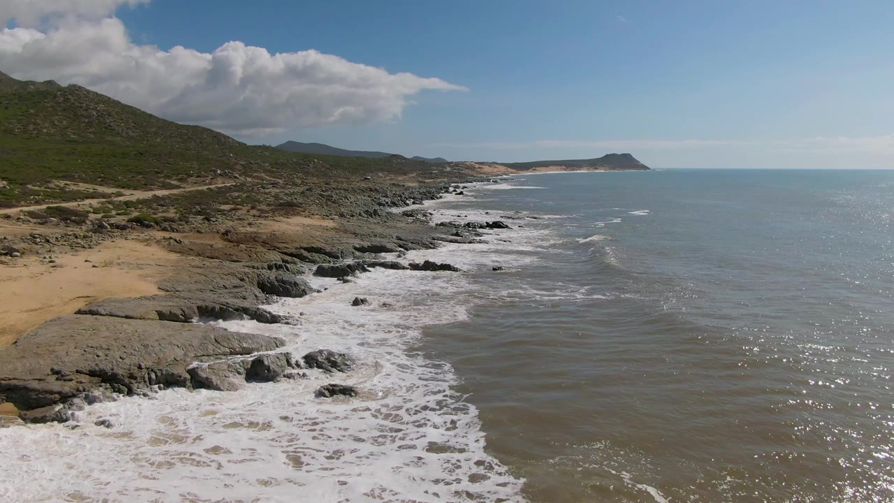 Mid-clip aerial view at Buzzards Beach East Cape BCS Mexico where the surf line thins along the rocks, the sandy beach remains at lower left, hills fill the left background, and no visible people appear.