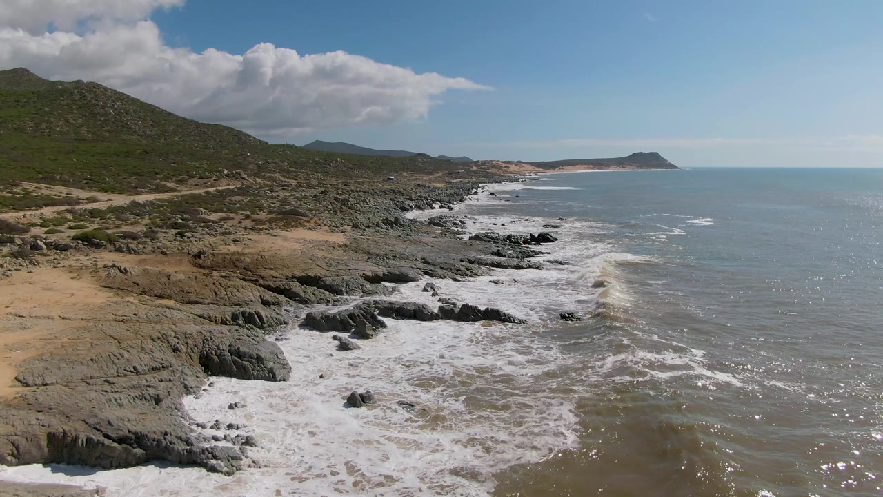 Rocky coastline with white surf, green hills, blue ocean, and partly cloudy sky.