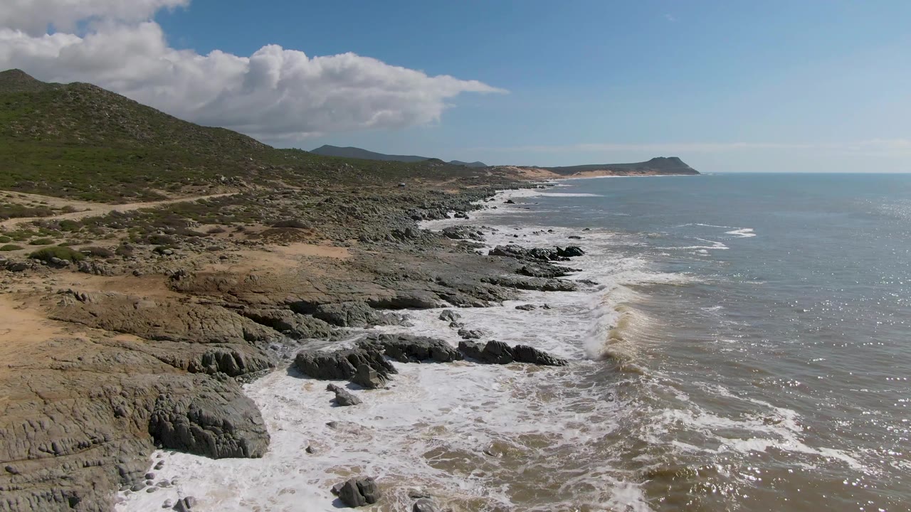 Aerial frame from Buzzards Beach East Cape in Mexico showing surf spreading farther across black rock shelves, tan shoreline on the left, open water on the right, and no visible people.