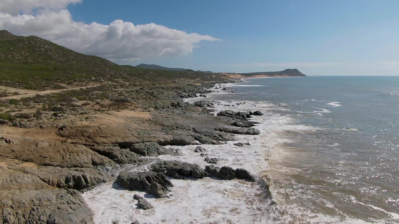 Aerial view at Buzzards Beach East Cape in Mexico with bright foam curling around foreground rocks, green hills rising inland, a distant headland near the horizon, and no visible people.
