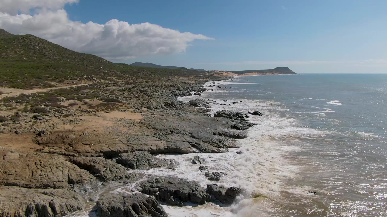 Aerial frame over Buzzards Beach East Cape in Mexico showing more tan sandy ground in the foreground, jagged rock ledges along the surf line, green hillside, blue water, and no visible people.