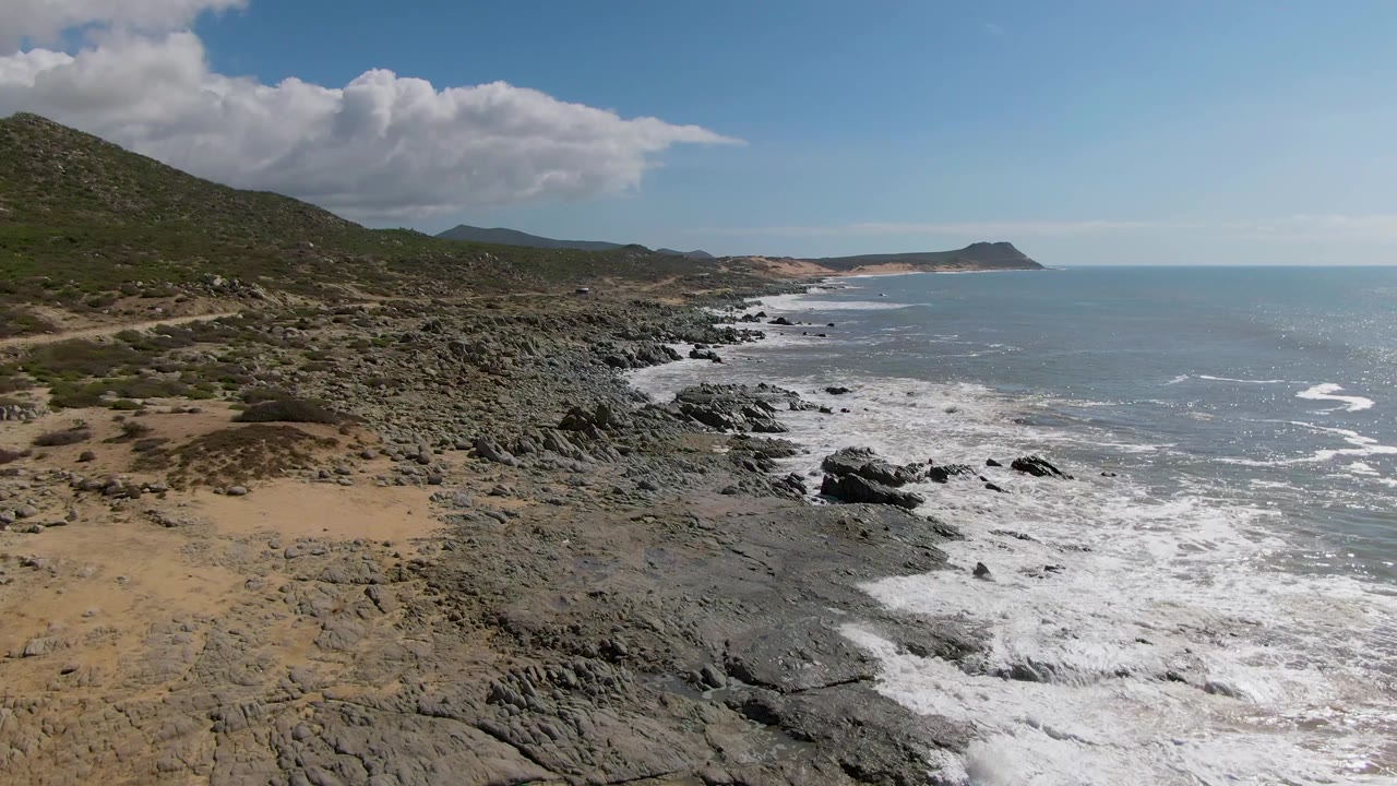 Wide aerial view of Buzzards Beach East Cape BCS Mexico with tan sand at left, dark rocky shore, white surf, green hills, distant headland, and no visible people.
