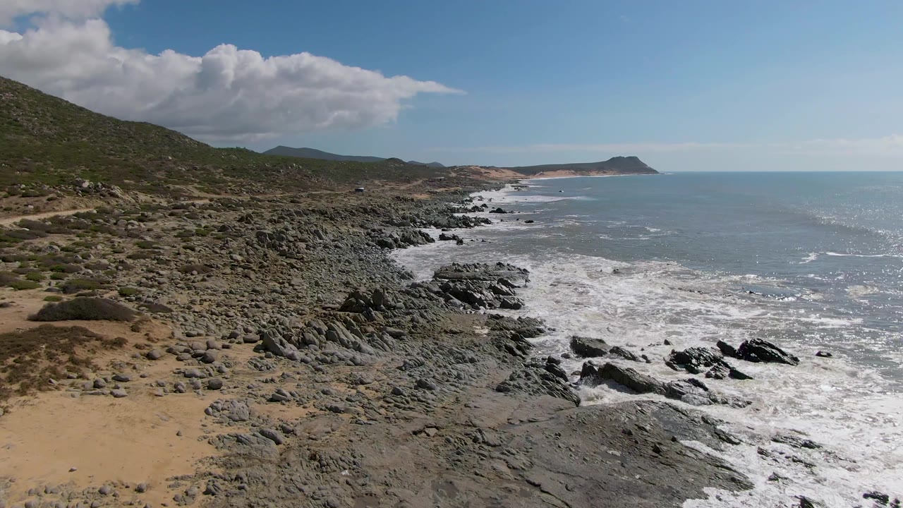 Buzzards Beach East Cape BCS Mexico coastline with rough boulders in the foreground, white water curling into the rocks, ocean to the right, and no visible people.