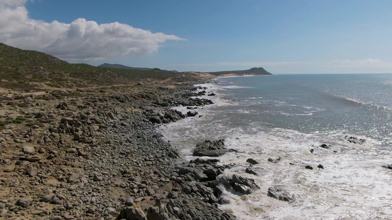 Elevated view of Buzzards Beach East Cape BCS Mexico with dark rocks, foamy surf, distant headland, hilly terrain, and no visible people at the opening moment.