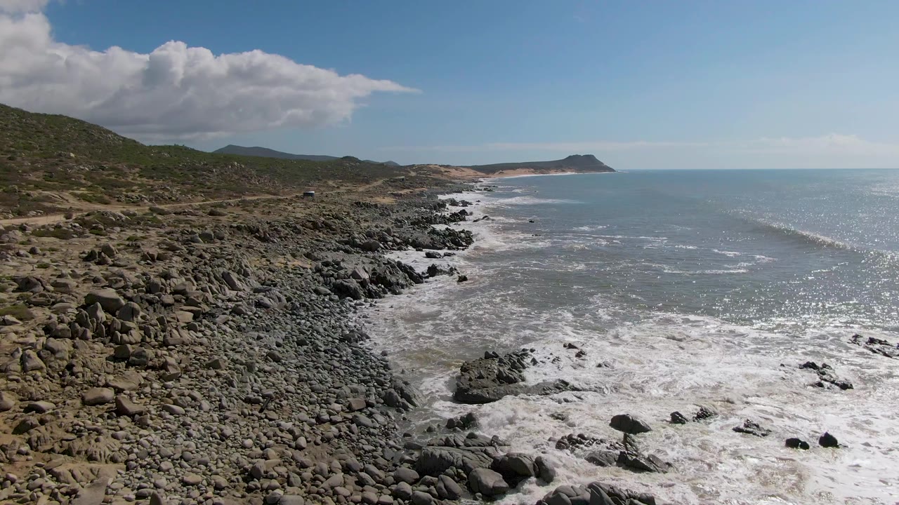 Buzzards Beach East Cape BCS Mexico shoreline seen from above as white water spreads across midground rocks, with scrubby hills on the left and no visible people.