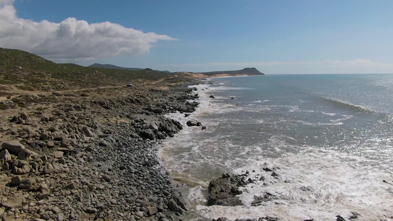 The Buzzards Beach East Cape BCS Mexico coast curves through frame with surf breaking over black rocks, bright sun glinting on the ocean, and no visible people.