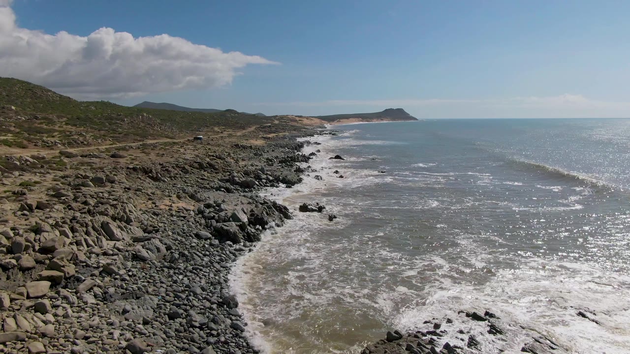 Mid-clip view of Buzzards Beach East Cape BCS Mexico with more open water on the right, rocky foreground shore on the left, rolling hills, and no visible people.