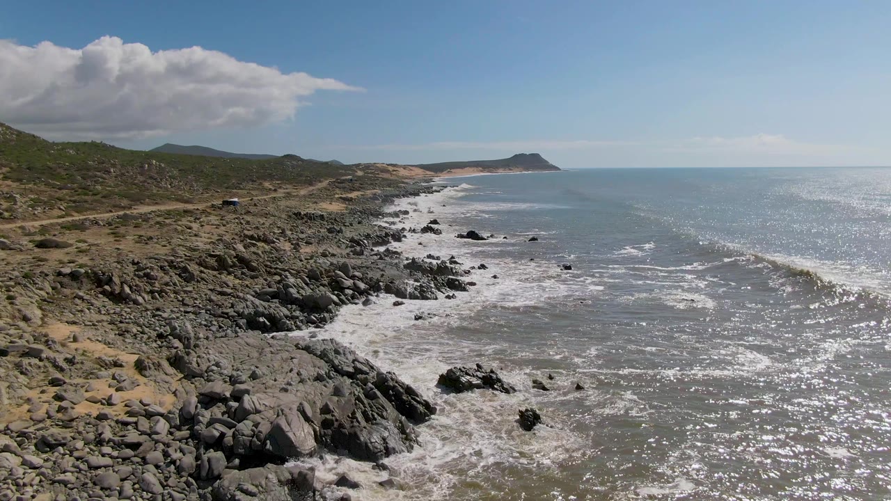 Aerial view of Buzzards Beach East Cape BCS Mexico with dark shoreline rocks, white surf, a distant road, open ocean, and no visible people at the start of the clip.
