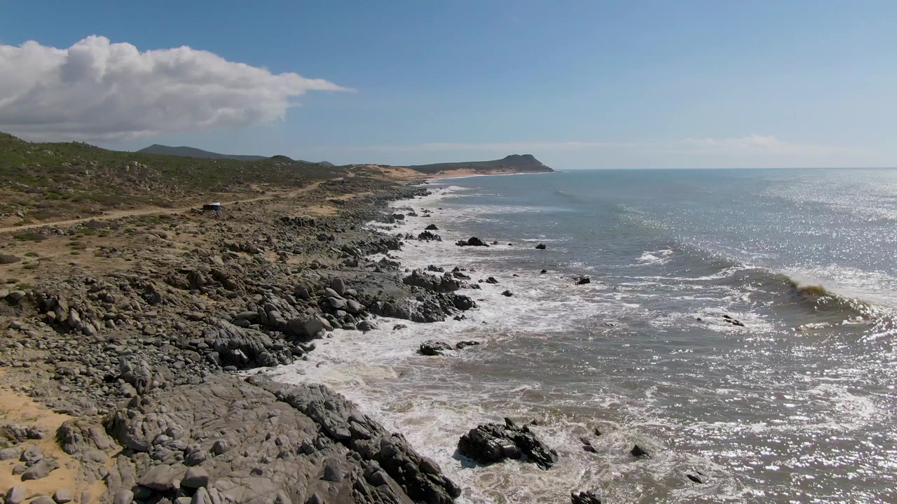 Buzzards Beach East Cape BCS Mexico coastline seen from above as foam spreads around nearshore rocks, with scrub land and a small distant vehicle along the road, and no visible people.