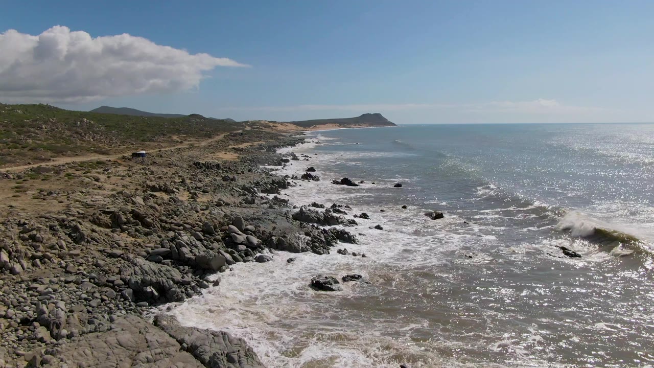 Aerial coastal frame at Buzzards Beach East Cape BCS Mexico with a broader sweep of surf over rocks, glinting ocean water to the right, distant headland, and no visible people.