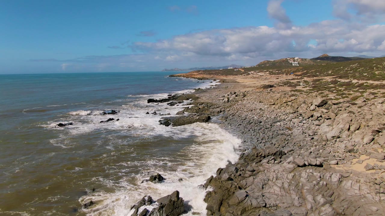 Aerial view of waves breaking on a rocky coast under scattered clouds.