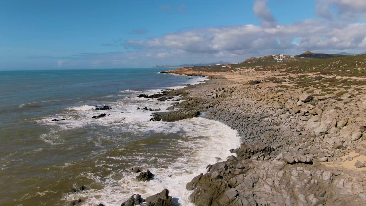 Aerial view at Buzzards Beach East Cape BCS Mexico with a larger band of foam spreading through the center rocks, open ocean to the left, rocky land to the right, and no visible people.