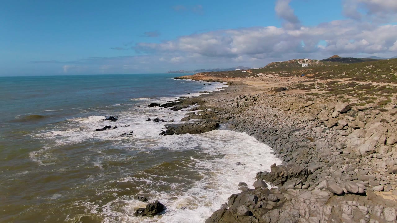 Aerial frame at Buzzards Beach East Cape BCS Mexico showing surf wrapped around black rock outcrops, a broad tan rocky shore on the right, distant hills, scattered clouds, and no visible people.