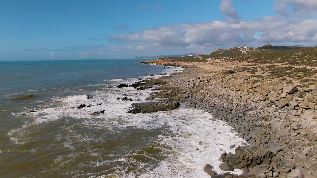 Aerial view at Buzzards Beach East Cape BCS Mexico with the wave line lower in frame, foamy water reaching the foreground rocks, open sea to the left, rocky coast ahead, and no visible people.