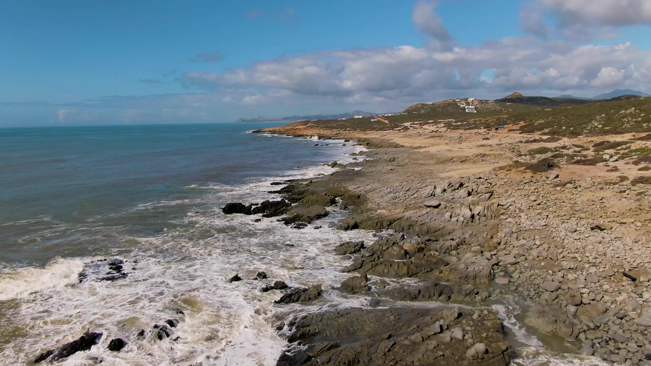 Drone-height coastal view of Buzzards Beach East Cape BCS Mexico with a larger wash of foam in the lower left, rocky shore through the center, distant headland, and no visible people.