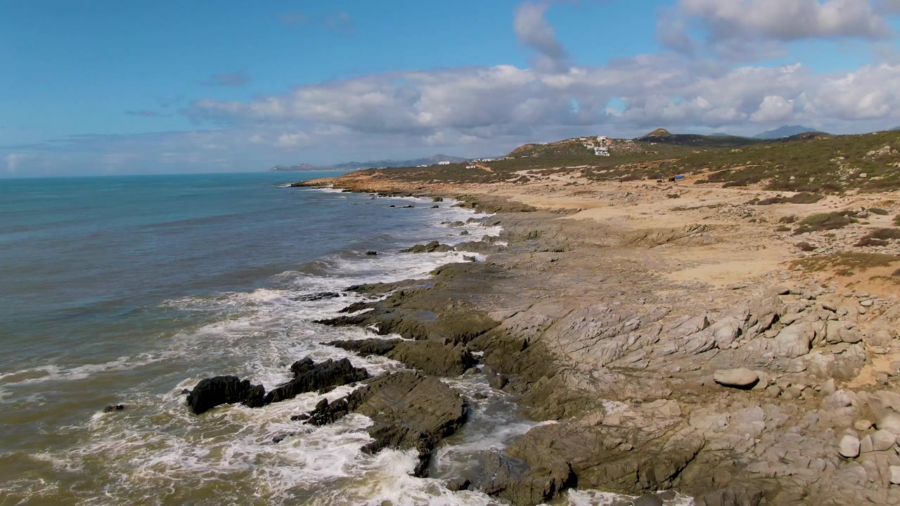 At Buzzards Beach East Cape BCS Mexico, waves break against dark rocks below a sandy beach and scrub-covered hills, with blue-green ocean to the left and no visible people.