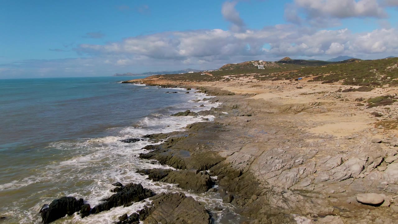 At Buzzards Beach East Cape BCS Mexico, white foam spreads across the foreground rocks while the sandy beach and low hills remain clear in the background, with no visible people.