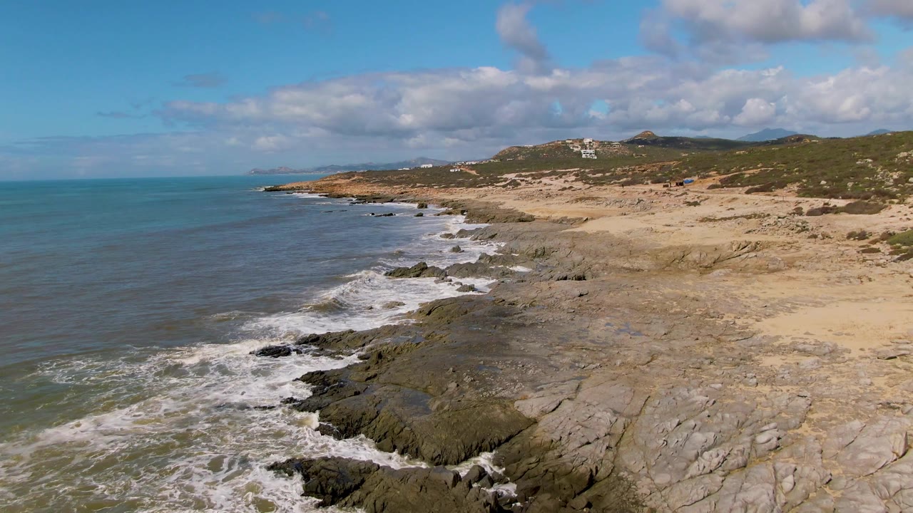 At Buzzards Beach East Cape BCS Mexico, the surf curls along the left side of the rocky shore while pale sand and green-brown hills stretch across the right, with no visible people.