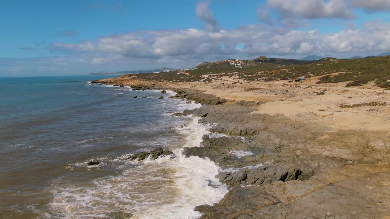 Aerial view of Buzzards Beach East Cape in Mexico with white waves breaking over dark rocks, tan sand to the right, blue-green ocean to the left, distant hillside buildings, and no visible people.