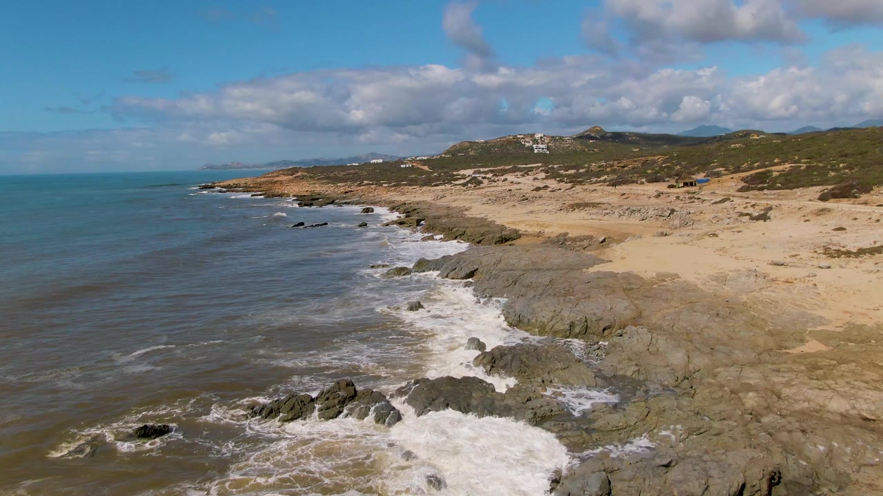 Aerial coastal frame at Buzzards Beach East Cape, Mexico, where foam spreads across the rocks in the foreground, the sandy beach runs inland, hills sit under clouds, and no visible people are present.