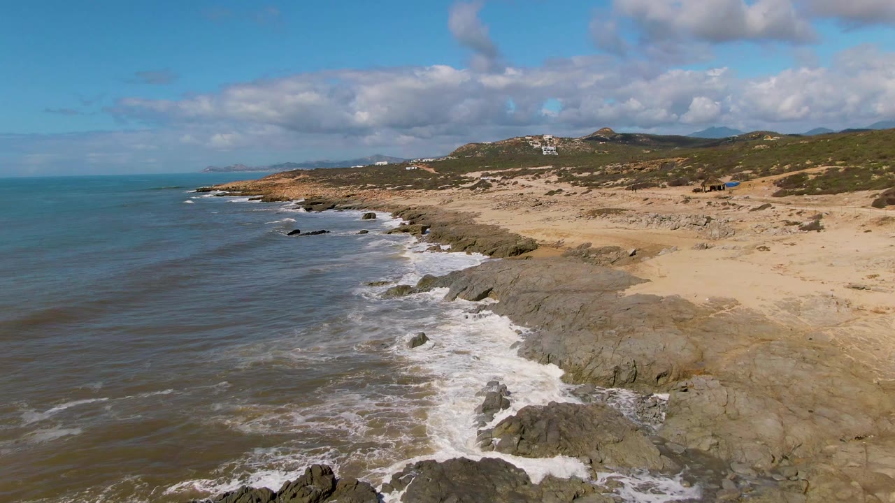 Buzzards Beach East Cape shoreline in Mexico with a longer surf line visible along the coast, dark rocks in the lower frame, tan beach to the right, partly cloudy sky above, and no visible people.