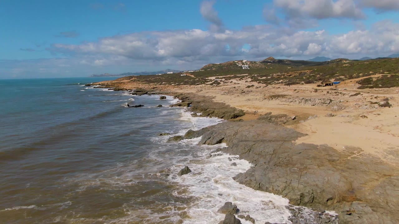 Waves wash across rocks below a sandy coast and hills