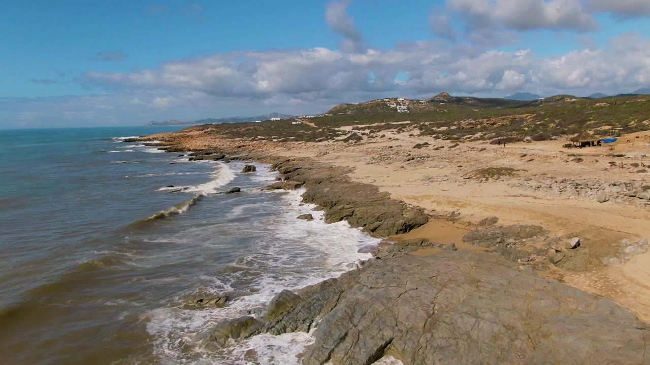 Rocky beach shoreline with waves and low hills under a partly cloudy sky