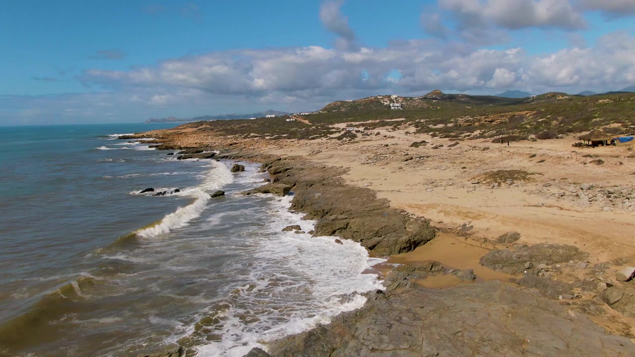 At Buzzards Beach East Cape BCS Mexico, a larger band of foam spreads over nearshore rocks while the sandy beach and scrubby hills remain clear in the background, with no visible people.