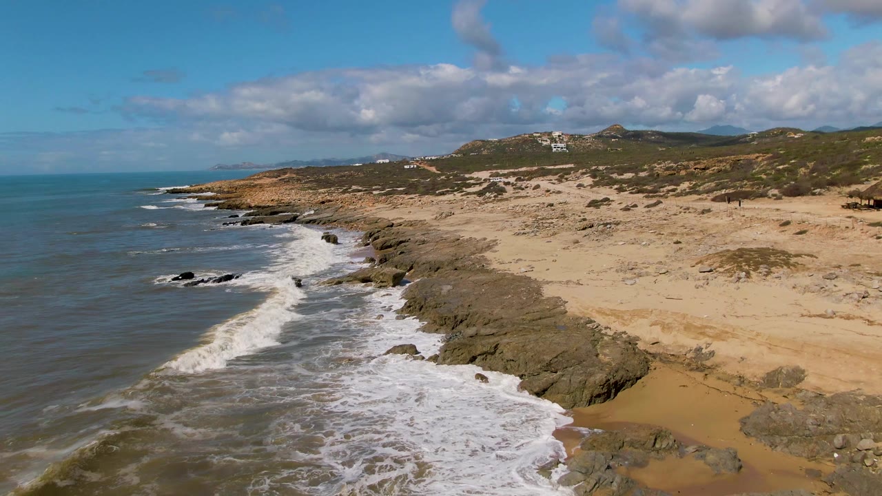 At Buzzards Beach East Cape BCS Mexico, white water curls along the rocky edge below a broad tan beach, with green-brown hills and scattered clouds behind it, and no visible people.