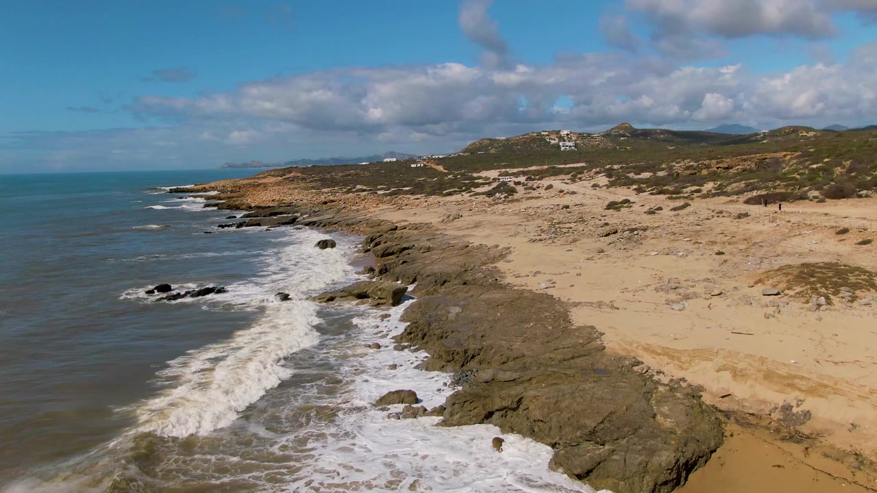 At Buzzards Beach East Cape BCS Mexico, the view shifts farther along the coast, showing a diagonal line of surf and rocks against open water, tan beach, and distant hills with no visible people.