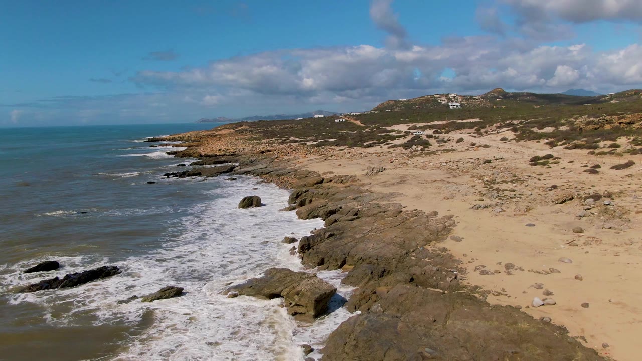 Aerial frame of Buzzards Beach East Cape in Mexico with surf washing over rocks, tan sand and dunes to the right, low hills in the distance, and no visible people.